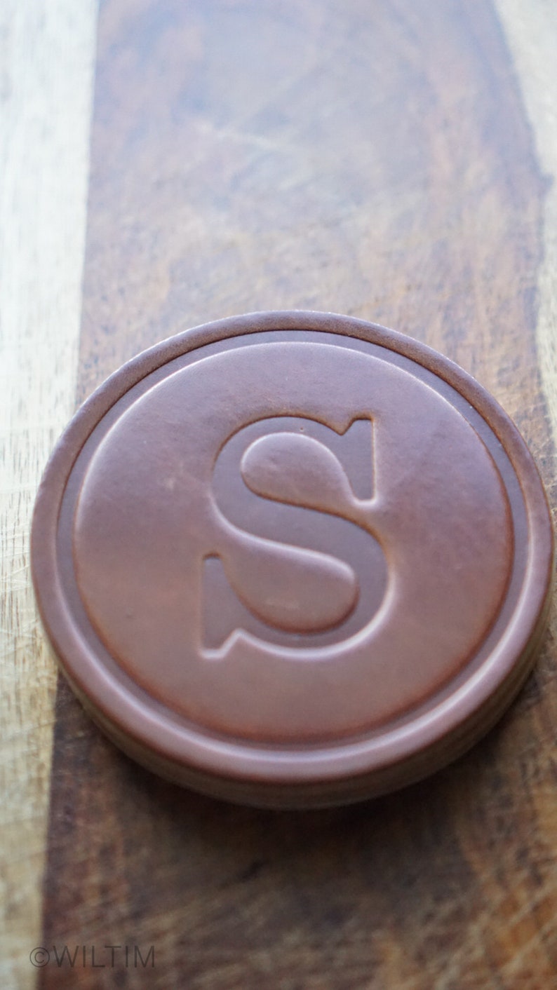 May include: A brown, circular coaster with a raised edge and a large, embossed letter "S" in the centre. The coaster is made of a solid material and rests on a wooden surface.