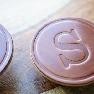 May include: Close-up of two brown leather coasters with an embossed letter "S" in the center. The coasters have a circular shape with a lighter-coloured edge. The background is a wooden surface.