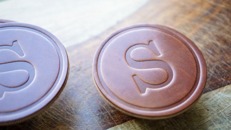May include: Close-up of two brown leather coasters with an embossed letter "S" in the center. The coasters have a circular shape with a lighter-coloured edge. The background is a wooden surface.