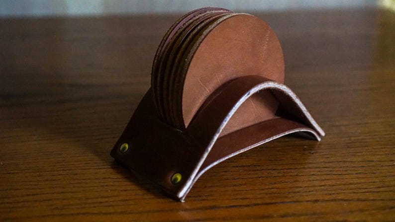 May include: A set of brown leather coasters and a holder. The coasters are round and stacked, with a curved holder. The holder has brass rivets. The leather has a natural, textured appearance.