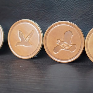 May include: Four brown leather coasters with different bird silhouettes embossed on them. The birds are a turkey, a duck, a quail, and a dove.