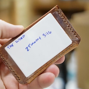 May include: A small, rectangular, brown leather card holder with a white card inside. The card has the words "The Word" and "2 Timothy 3:16" written in blue ink. The holder is held in a person's hand.