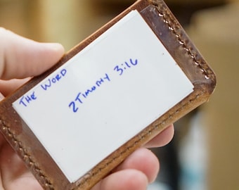 May include: A small, rectangular, brown leather card holder with a white card inside. The card has the words "The Word" and "2 Timothy 3:16" written in blue ink. The holder is held in a person's hand.