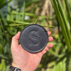 May include: A black, circular drinks coaster featuring a debossed "S" in the centre. The coaster is held in a hand, with a black watch on the wrist. Green foliage forms the background.