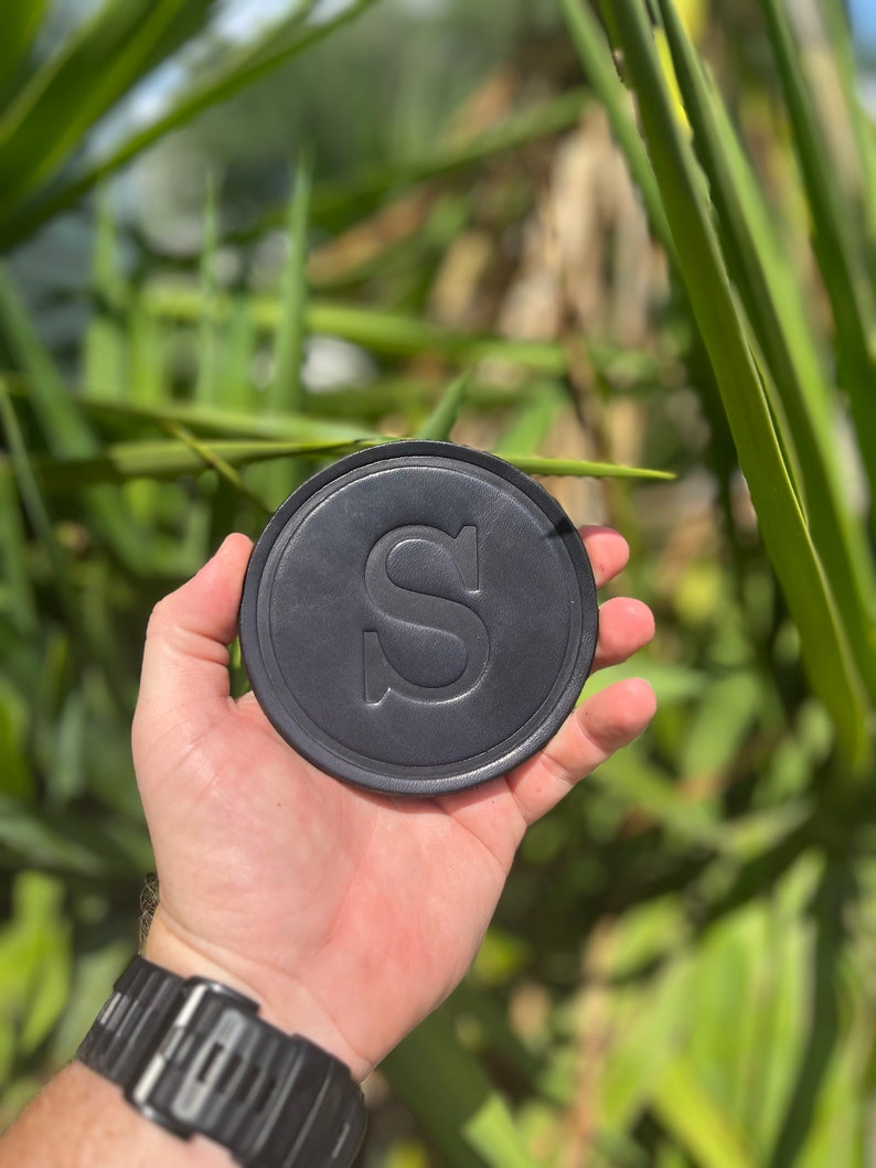 May include: A black, circular drinks coaster featuring a debossed "S" in the centre. The coaster is held in a hand, with a black watch on the wrist. Green foliage forms the background.