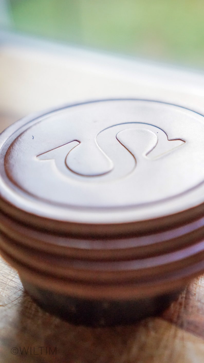 May include: Close-up of a brown, circular object with a white, stylized "S" design on top. The object has several stacked, ridged layers. The background is blurred, with a wooden surface in the foreground.