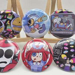 May include: Six circular pin-back buttons featuring colorful cartoon illustrations. Designs include a yellow face with geometric patterns, a blue-toned character holding a cup, a jester on a television screen, a crying face, a red-haired doll, and a dark background with multiple eyes and computer error text.