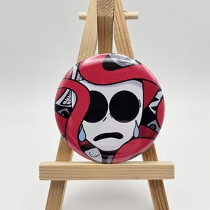 May include: A circular pinback button featuring a stylized white face with large black eyes and blue teardrops, surrounded by thick red and black abstract shapes, displayed on a small wooden easel stand for clear visibility and presentation.
