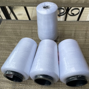 May include: Four spools of white thread wrapped in clear plastic. The spools are arranged in a row on a brown woven surface. The spools are labeled "Made in China".