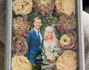 May include: A framed photo of a couple in a wedding setting, surrounded by dried flowers in shades of beige, brown, and purple. The flowers are arranged in a circle around the photo, creating a decorative border.