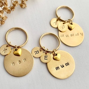 May include: Three gold-toned metal keychains, each featuring a large circular disc stamped with the words nan, mum, or mummy. Each keychain includes smaller circular discs stamped with letters K, R, E, A, G, and a small gold-toned heart charm.