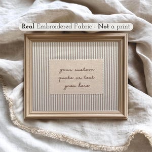 May include: A rectangular light wood frame containing a piece of beige fabric with vertical white stripes. Centered on the fabric is a rectangular patch of off-white fabric with pinked edges, featuring the embroidered text: your custom quote or text goes here.
