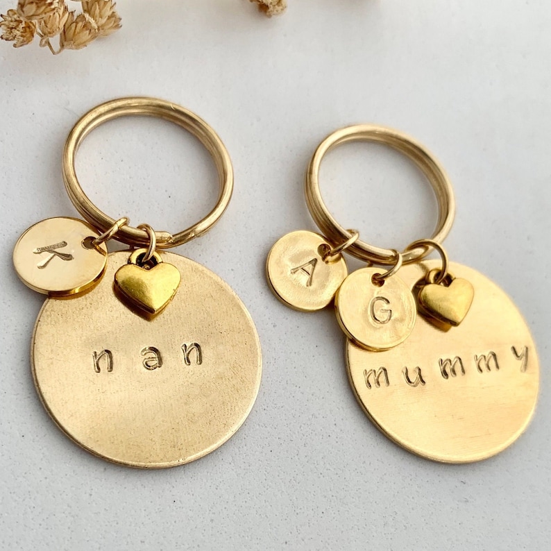May include: Two gold-toned metal keychains featuring circular discs. The left keychain is stamped with the word nan and a small disc with the letter K. The right keychain is stamped with the word mummy and two small discs with the letters A and G.
