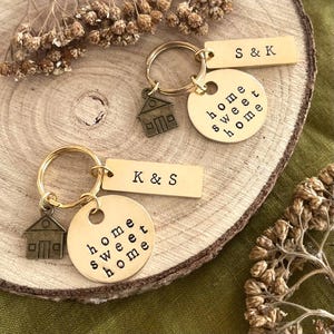 Puede incluir: Dos llaveros de metal dorado, cada uno con un disco circular grabado con el texto home sweet home, una etiqueta rectangular con las iniciales S & K o K & S, y un pequeño dije en forma de casa de color bronce en una anilla.