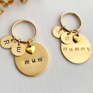 May include: Two gold-colored metal keychains featuring circular discs. One keychain has a large disc stamped with the word mum, two smaller discs stamped with R and E, and a heart charm. The other has a large disc stamped with mummy, two smaller discs stamped with A and G, and a heart charm.