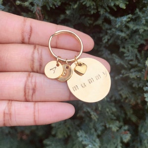 May include: A gold-toned metal keychain featuring a large circular disc engraved with the word mummy, a small heart-shaped charm, and two smaller circular discs, one of which is stamped with the capital letter A, all attached to a split ring.