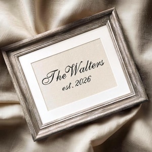 May include: A rectangular picture frame with a weathered grey wood finish and a white mat. Inside the frame, a cream-coloured fabric insert features black embroidered text that reads The Walters and below it, the text est. 2026 in a cursive font.