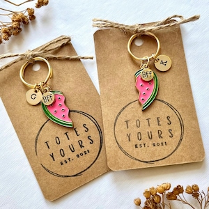 Puede incluir: Dos llaveros dorados con dijes de rebanadas de sandía, la palabra "BFF" y dijes con iniciales. Los llaveros están sujetos a tarjetas marrones con el texto "TOTES YOURS EST. 2021". Los dijes de sandía son rosas, verdes y negros.