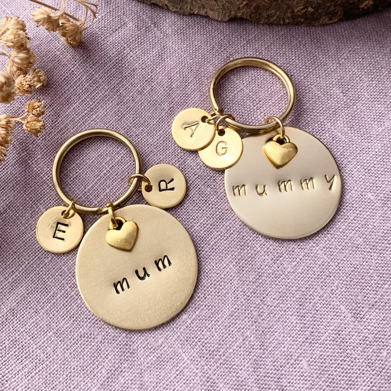 May include: Two gold-toned metal keychains featuring circular discs. One keychain is stamped with the word mum and the letter E and R. The other keychain is stamped with the word mummy and the letters A and G. Each includes a small heart charm.