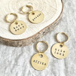 Puede incluir: Cuatro llaveros dorados con etiquetas redondas. Cada etiqueta tiene una palabra diferente estampada: "back door", "garage", "office" y "bike lock". Los llaveros están sobre una superficie de madera.