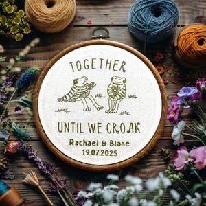 Op de afbeelding: Een ronde houten borduurring met witte stof en groen geborduurde tekst TOGETHER UNTIL WE CROAK, Rachael & Blane en 19.07.2025, voorzien van twee geborduurde kikkers, waarvan er één een trui draagt en de andere een bloem vasthoudt.