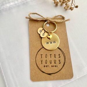 May include: A gold-toned metal keychain featuring a circular disc stamped with the word mum, a small heart charm, and two smaller discs stamped with the letters R and E. The keychain is attached to a rectangular kraft paper tag printed with TOTES YOURS EST. 2021.