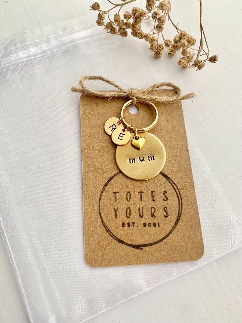 May include: A gold-toned metal keychain featuring a circular disc stamped with the word mum, a small heart charm, and two smaller discs stamped with the letters R and E. The keychain is attached to a rectangular kraft paper tag printed with TOTES YOURS EST. 2021.