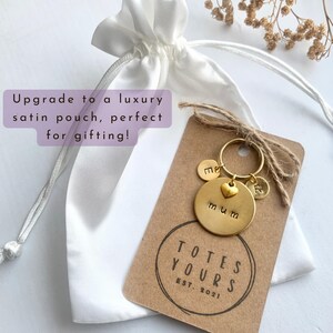 May include: A gold-toned metal keychain featuring a large circular disc stamped with the word mum, a small heart charm, and two smaller circular discs stamped with the letters E and R. The keychain is attached to a brown card labeled TOTES YOURS EST. 2021.