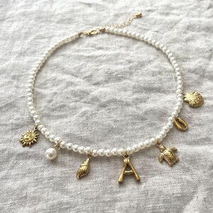 Könnte beinhalten: Eine Perlenkette mit goldfarbenen Anhängern. Die Anhänger umfassen eine Sonne, eine Perle, eine Muschel, den Buchstaben "A", eine Schildkröte und eine Schnecke. Die Kette liegt auf einer weißen Leinenoberfläche.