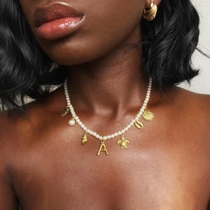 Puede incluir: Un collar de perlas con colgantes dorados, incluyendo una concha, una tortuga y la letra "A". El collar se lleva alrededor del cuello. Los colgantes son dorados y las perlas son blancas.