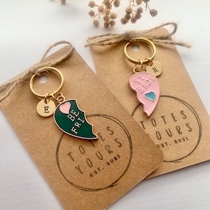 May include: Two gold keychains with broken heart charms. One charm is green with the text "Be Friends" and the other is pink with the text "Best Friends". Both charms have a small heart on them. The keychains are on brown card stock with the text "Totes Yours Est. 2021" printed on them.