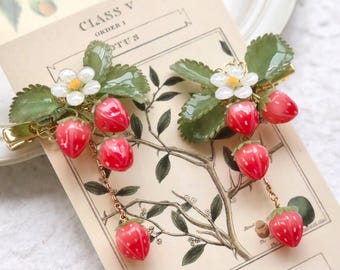 May include: Two gold hair clips with a strawberry and flower design. Each clip has a chain with a dangling strawberry. The clips are on a vintage botanical illustration with the text "CLASS V ORDER I OTUS NUS LOTUS".