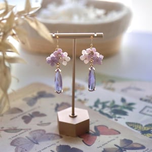 May include: A pair of dangle earrings on a gold stand. Each earring has a cluster of light purple and white flower beads, with a teardrop-shaped, light purple crystal pendant. The earrings are on a gold hook. The stand is on a patterned surface.