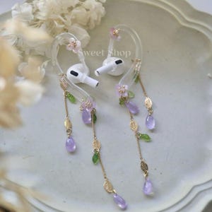 Könnte beinhalten: Weiße kabellose Ohrhörer mit transparenten Ohrhaken, verziert mit rosa Blüten, goldenen Blättern und hängenden lila tropfenförmigen Perlen. Der Text "Sweet Shop" ist sichtbar. Die Ohrhörer liegen auf einem hellen Teller.