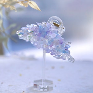May include: A clear hair claw clip, decorated with blue and purple flower-shaped beads, resembling hydrangeas. The clip is made of clear plastic and is displayed on a clear stand. The beads have a delicate, shimmering appearance, adding to the clip's charm.