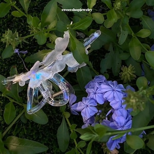 Könnte beinhalten: Eine transparente, schillernde Schmetterlingshaarspange mit einer silbernen Feder. Die Spange liegt auf einem Bett aus grünem Moos und violetten Blumen.