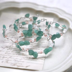 Puede incluir: Tres pulseras de alambre plateado con cuentas verdes en forma de hoja y pequeñas cuentas blancas y rosadas.
