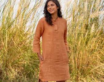 May include: A woman wearing a rust-colored linen dress with long sleeves and a mandarin collar. The dress has pockets and is paired with black ankle boots. The background features tall, dry grasses.
