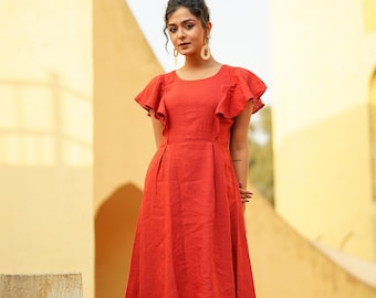 May include: A woman wearing a red linen dress with ruffled sleeves. The dress has a fitted bodice and a flowing skirt. She is standing on a stone platform in front of a yellow building.