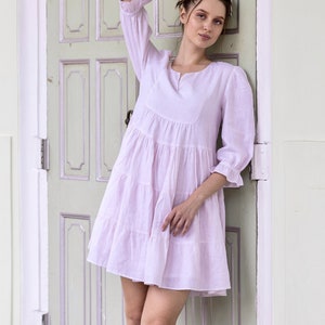 May include: A woman wearing a light pink tiered dress with a V-neck and 3/4 sleeves. She is standing in front of a light green door with white trim.