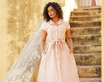 May include: A woman wearing a light pink linen dress with a button-down front and a tie belt. She is standing on a set of stairs with a yellow wall behind her.