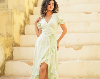 May include: A woman wearing a light green wrap dress with a ruffled hem and white sandals stands on a set of concrete stairs.