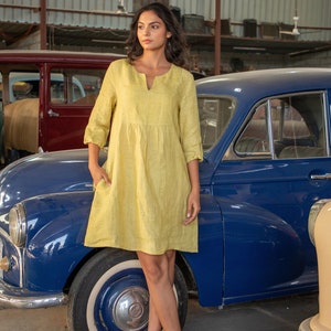 May include: A woman wearing a yellow linen dress stands in front of a blue vintage car. She is wearing white sandals and has her hair pulled back.