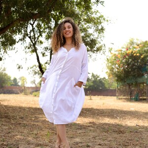 May include: A woman with curly hair is wearing a white, long-sleeved, button-down dress with pockets. She is standing in a field with green grass and trees in the background.