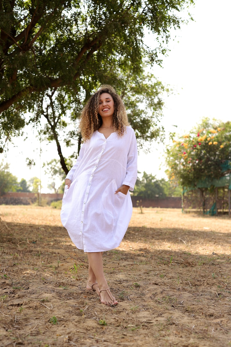 May include: A woman with curly hair is wearing a white, long-sleeved, button-down dress with pockets. She is standing in a field with green grass and trees in the background.