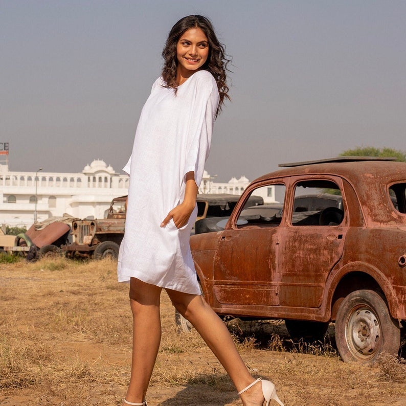 Puede incluir: Una mujer con un vestido blanco de lino est&aacute; de pie frente a un coche vintage oxidado. Ella sonr&iacute;e y mira hacia la derecha. El coche est&aacute; estacionado en un campo con un edificio blanco en el fondo.