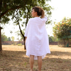 May include: A woman wearing a white linen dress stands in a field with a tree in the background.