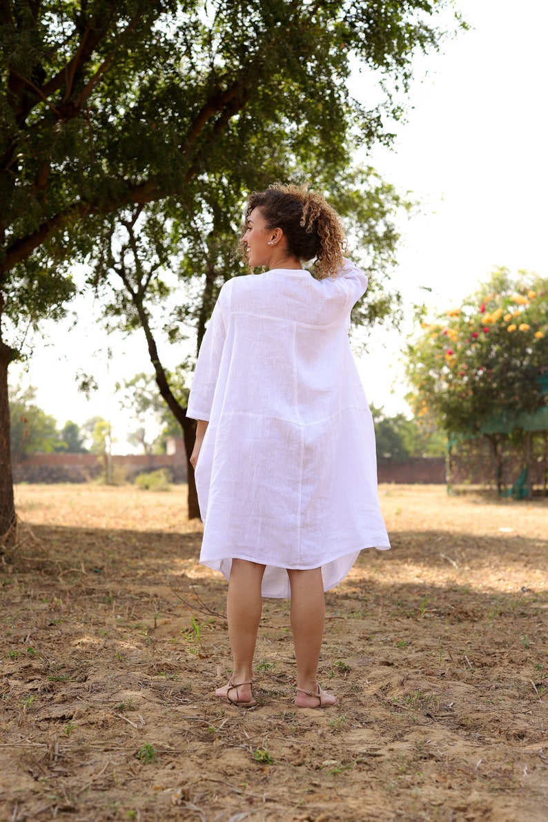 May include: A woman wearing a white linen dress stands in a field with a tree in the background.