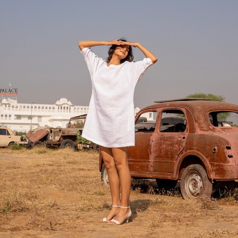 Puede incluir: Una mujer con un vestido blanco de lino est&aacute; de pie frente a un coche oxidado. Se est&aacute; protegiendo los ojos del sol con las manos. El coche est&aacute; aparcado en un campo con hierba seca.
