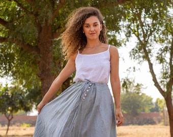 May include: A woman with curly brown hair wearing a white tank top and a gray linen skirt. She is standing in a field with trees in the background.